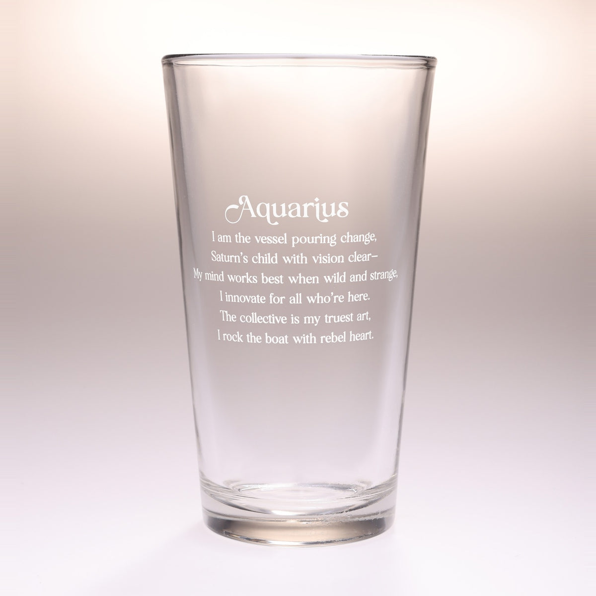 A clear glass drinking tumbler with etched Aquarius zodiac text and quote on the front, photographed against a soft beige-to-white gradient background.