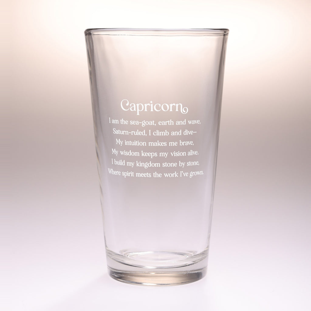 A clear glass pint glass with frosted white text reading 'Capricorn' and a poem underneath, shown on a soft light beige gradient background.