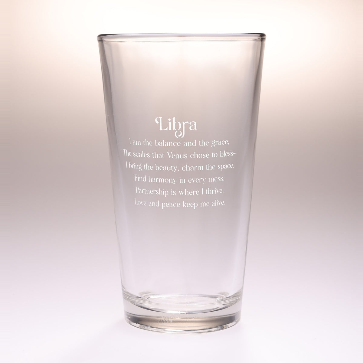 A clear glass drinking tumbler with etched white text reading 'Libra' and a poem associated with Libra, shown on a soft light gray to white gradient background.
