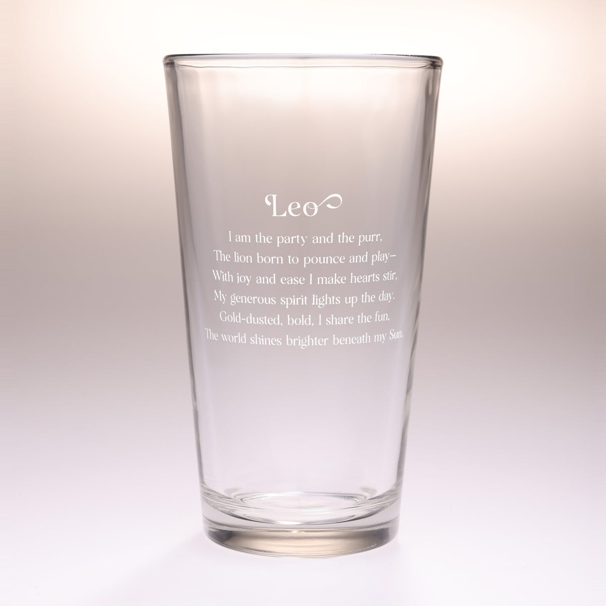 A clear glass drinking tumbler with etched Leo zodiac-themed text on the front, shown against a soft beige gradient background.