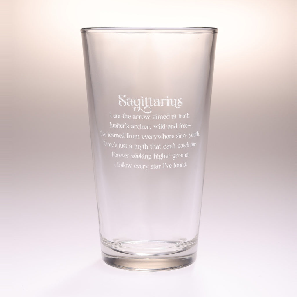 A clear glass pint-style drinking cup with etched white text reading 'Sagittarius' and a descriptive astrological quote, shown on a soft light beige-to-white studio background.