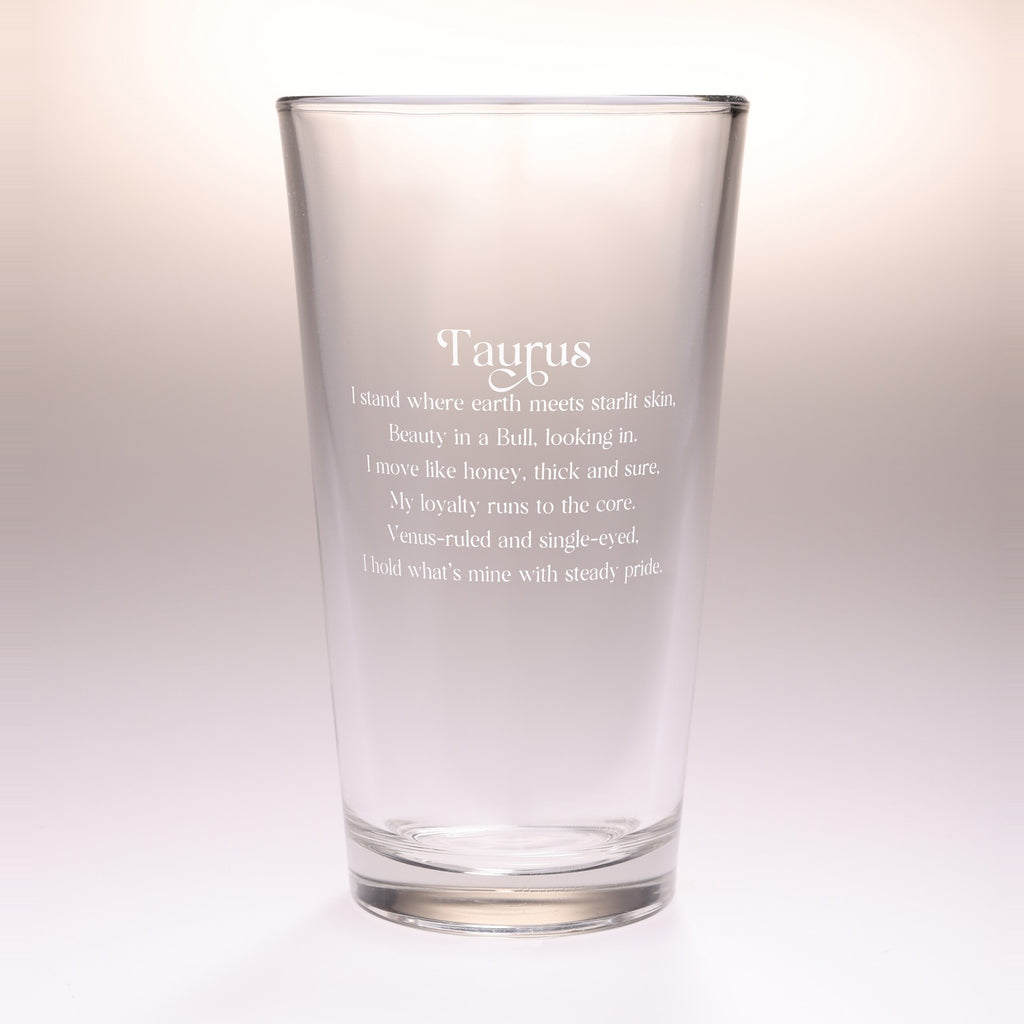 A clear glass drinking tumbler with etched white text reading 'Taurus' and a poem beneath it, shown on a soft light beige studio background.