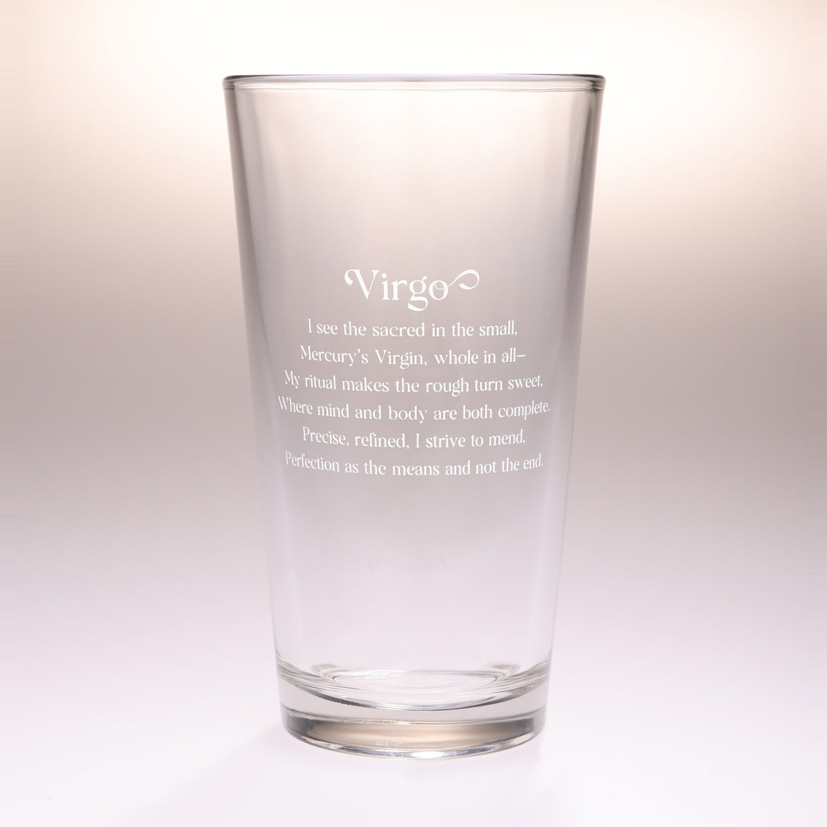 A clear glass drinking tumbler with etched white text reading 'Virgo' and a poem below, photographed against a soft light beige-to-white gradient background.