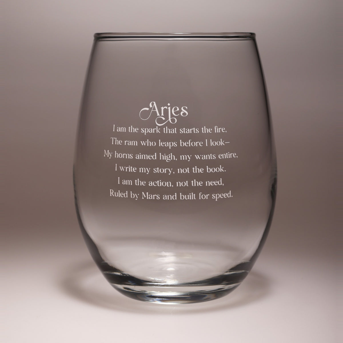A clear stemless wine glass with white engraved text reading 'Aries' and a poem about the zodiac sign, shown on a neutral gray background.