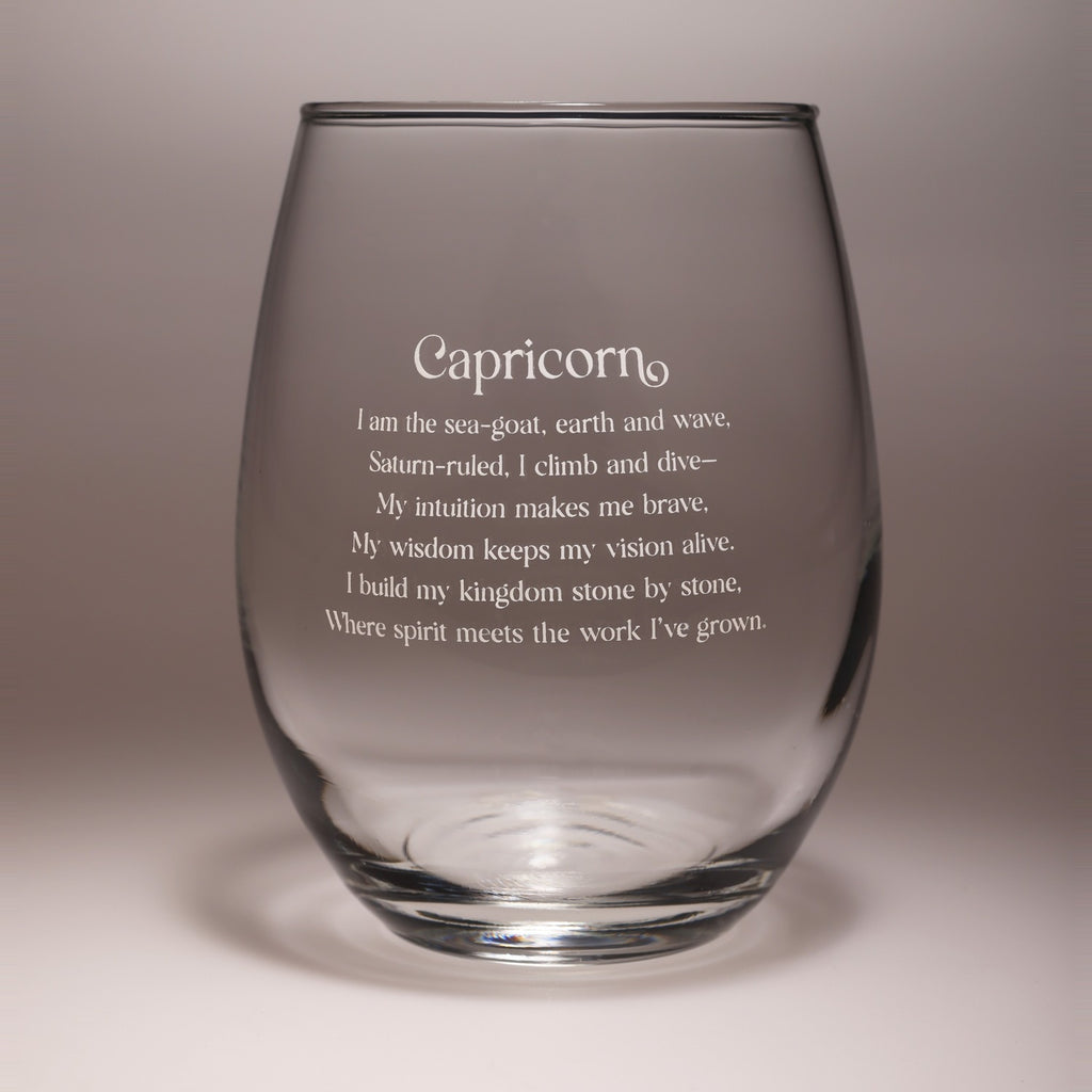 A clear stemless wine glass with etched Capricorn zodiac text on the front, shown against a neutral gray studio background.