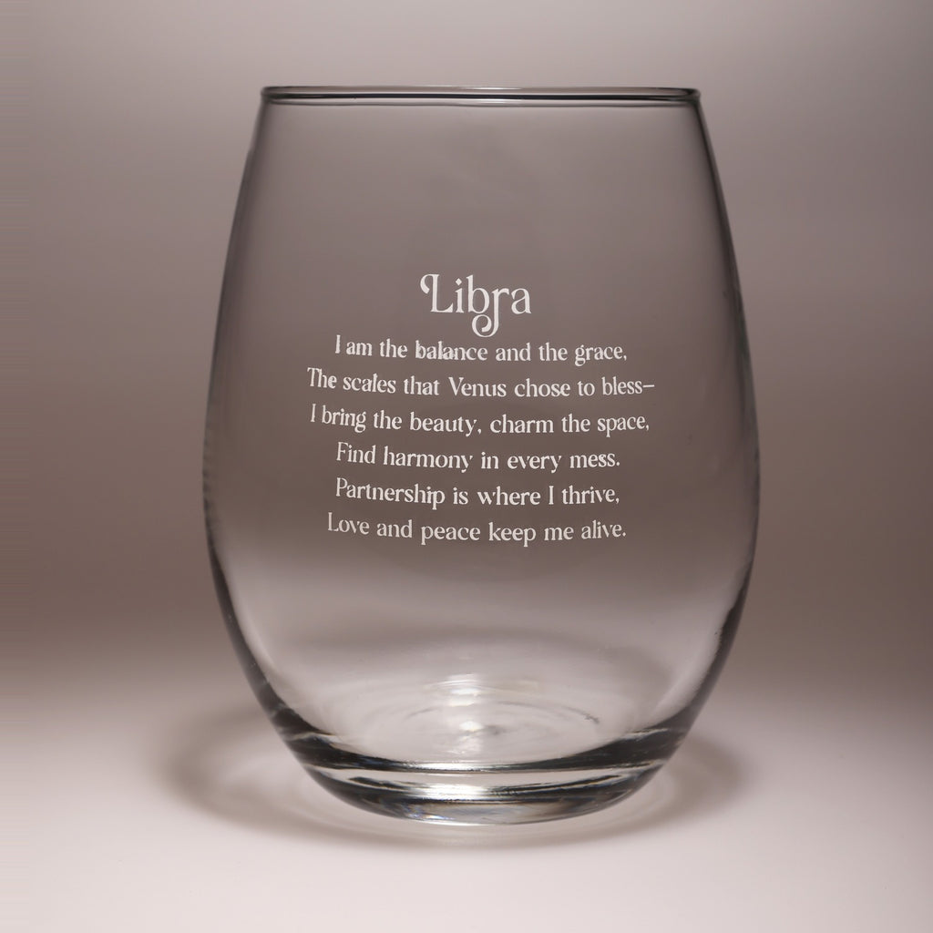 A clear stemless wine glass with frosted white text that reads 'Libra' followed by a poem about balance, grace, beauty, and harmony, shown on a neutral gray background.