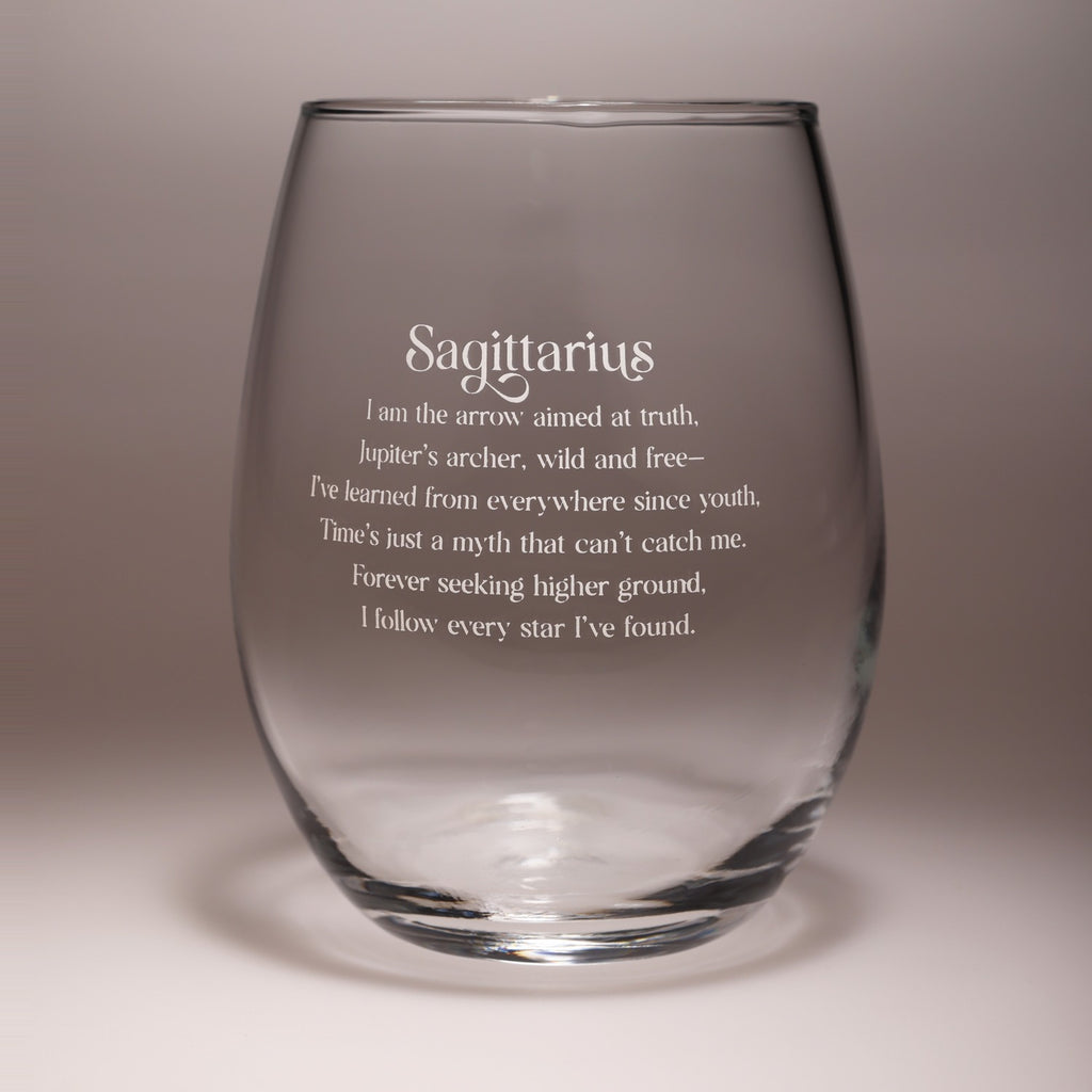 A clear stemless wine glass with frosted white text that reads 'Sagittarius' followed by a poetic description, shown on a neutral gray background.