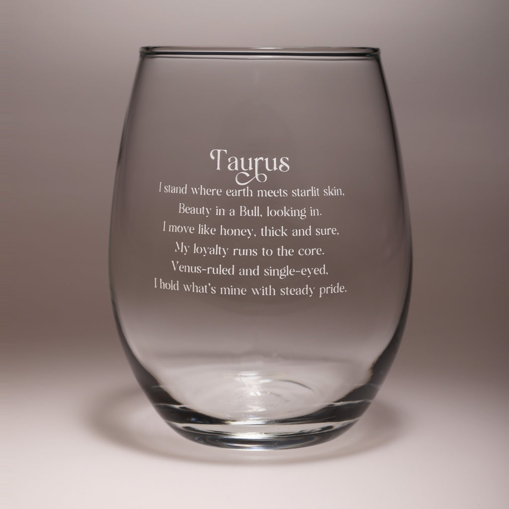 A clear stemless wine glass with white printed text reading 'Taurus' and a poem about the zodiac sign, shown on a neutral gray background.