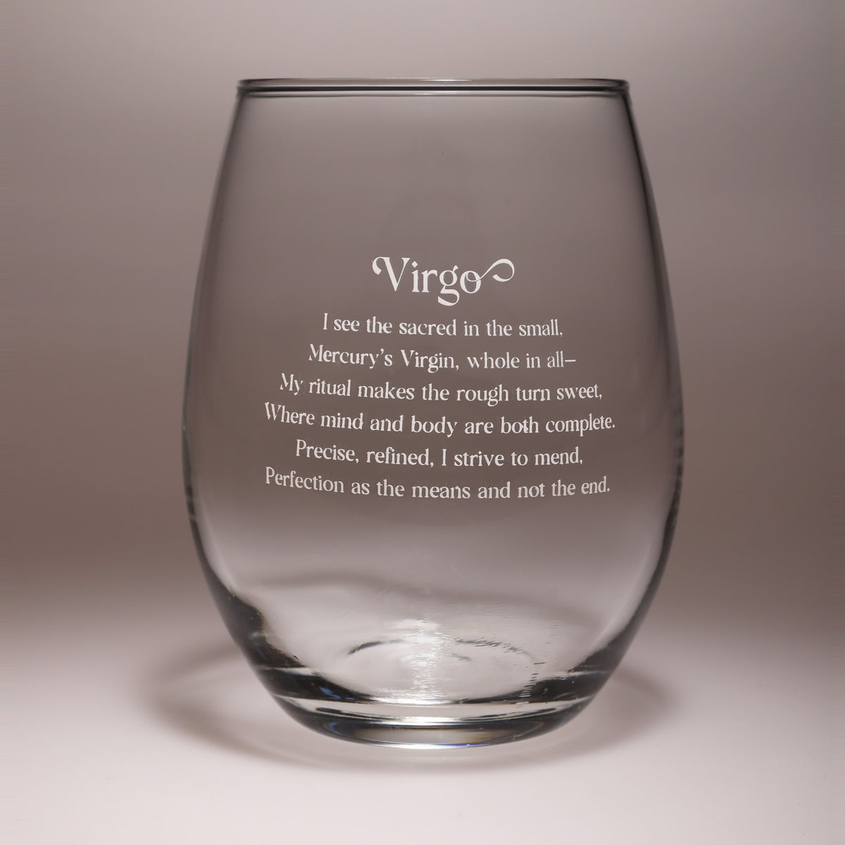 A clear stemless wine glass with white engraved text featuring the word 'Virgo' and a short poem, shown on a neutral gray background.