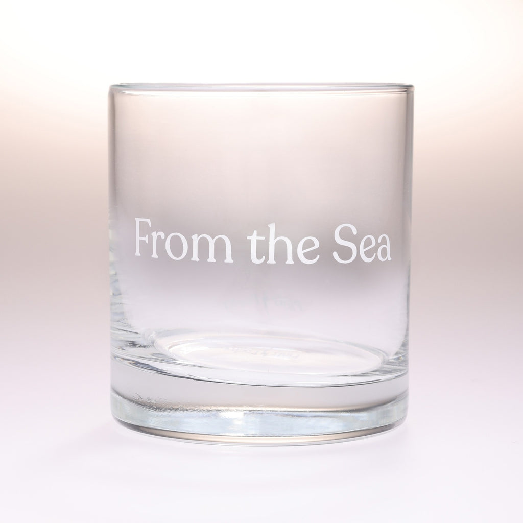 A clear glass rocks tumbler with the phrase 'From the Sea' printed in white serif lettering on the front, set against a soft white background.