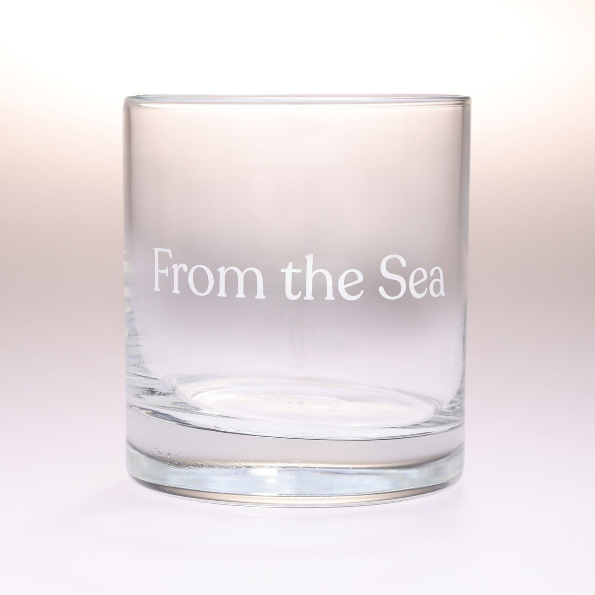 A clear glass rocks tumbler with the phrase 'From the Sea' printed in white serif lettering on the front, set against a soft white background.