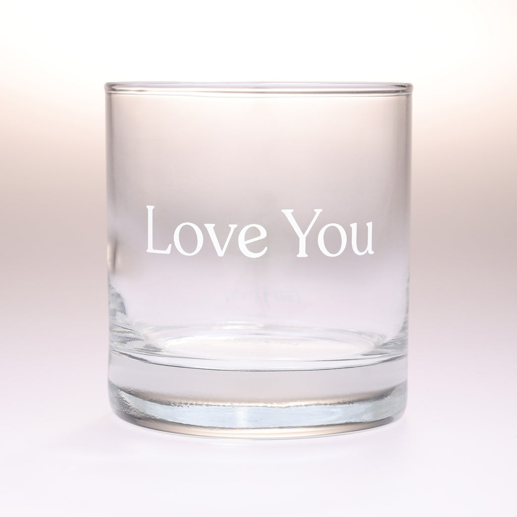 A clear glass rocks tumbler with the words 'Love You' printed in white serif lettering on the front, set against a soft white background.