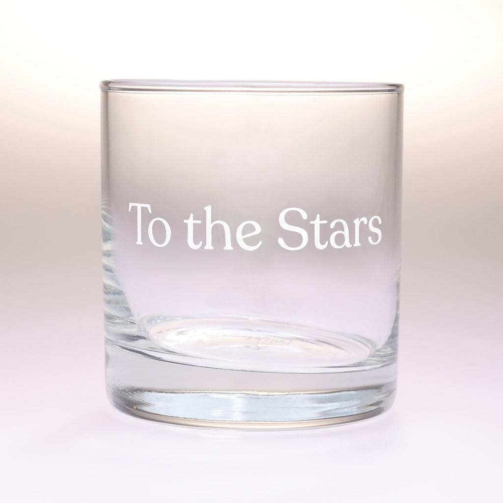 A clear glass rocks tumbler with the phrase 'To the Stars' printed in white serif lettering on the front, set against a soft white background.