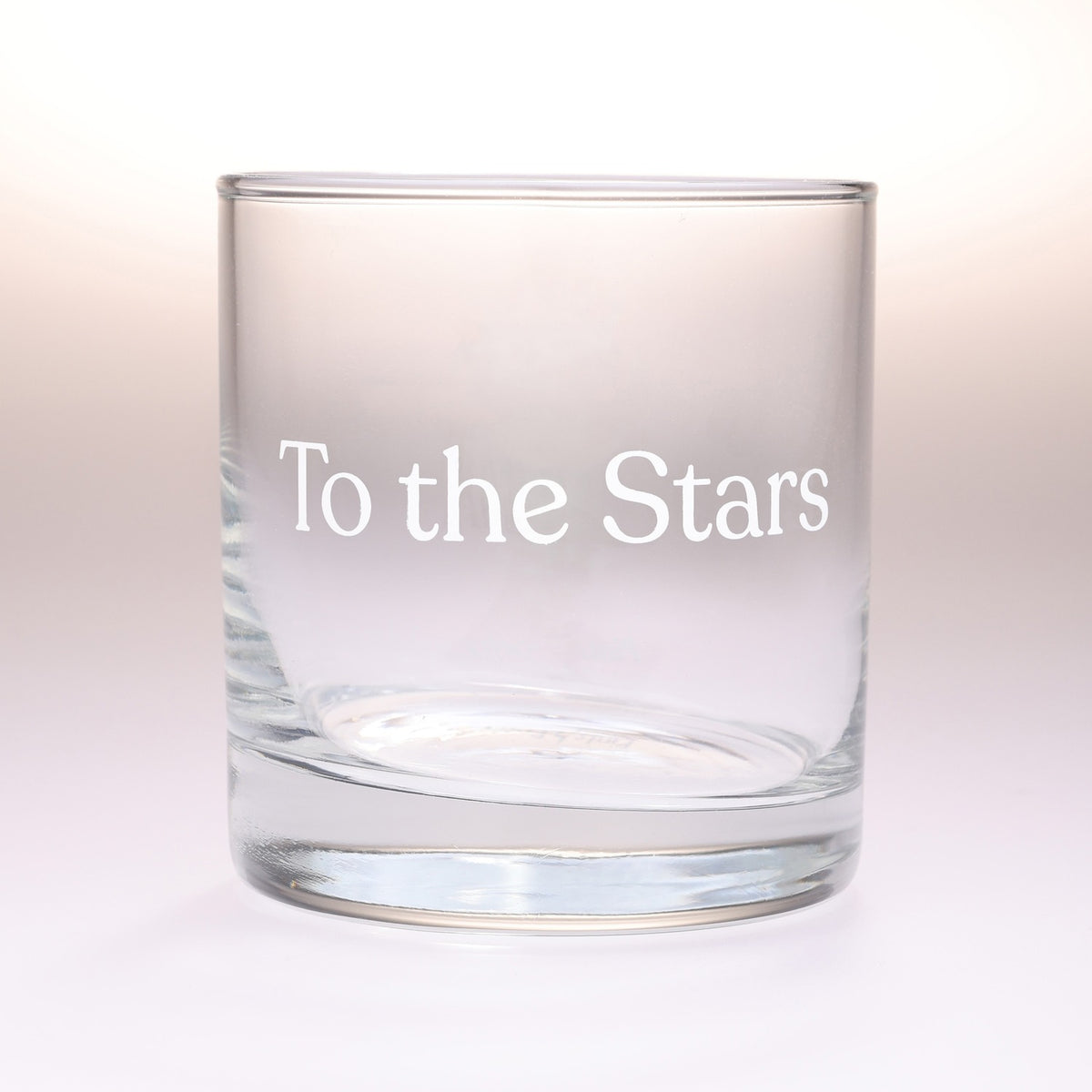 A clear glass rocks tumbler with the phrase 'To the Stars' printed in white serif lettering on the front, set against a soft white background.
