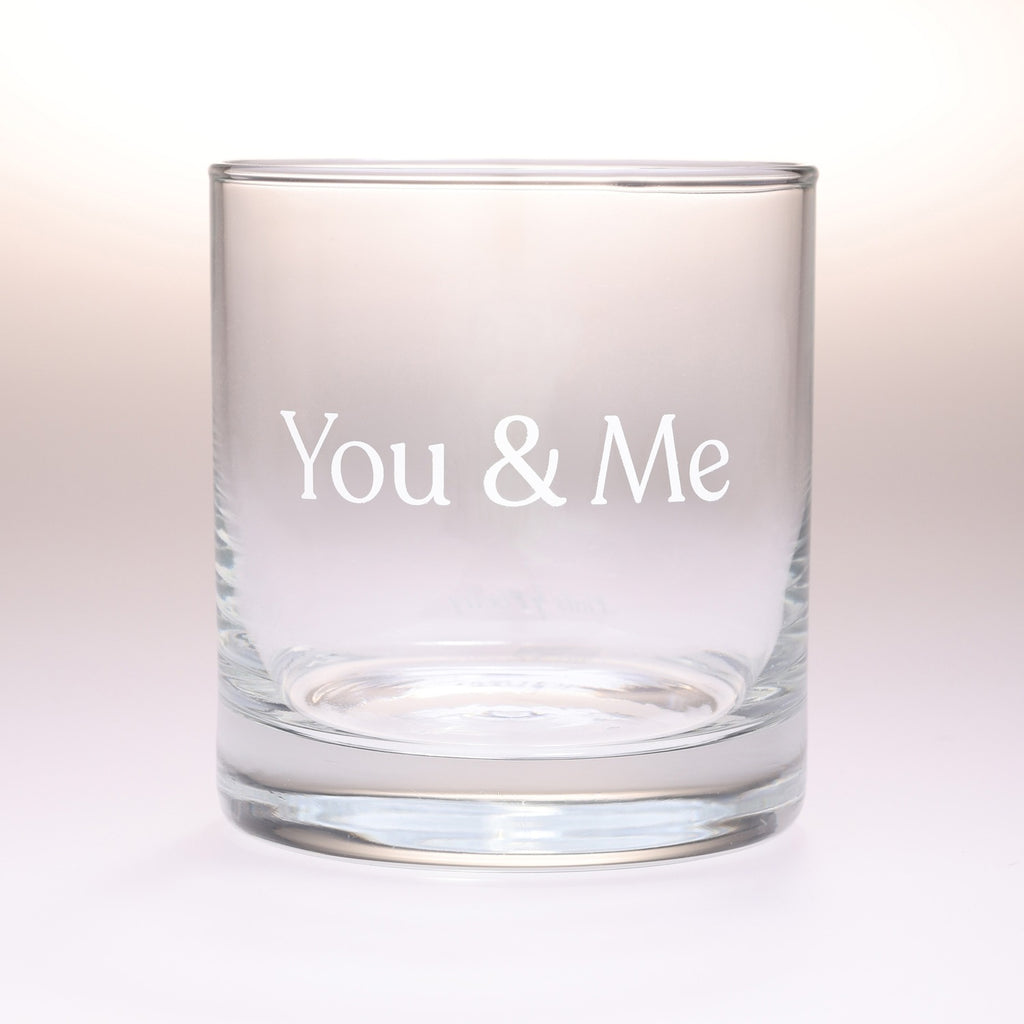 A clear glass rocks tumbler with the words 'You & Me' printed in white serif lettering on the front, set against a soft, light beige gradient background.