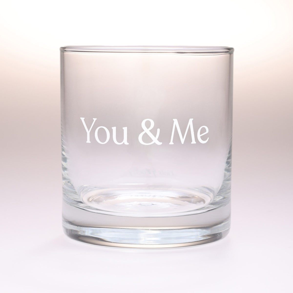 A clear glass rocks tumbler with the words 'You & Me' printed in white serif lettering on the front, set against a soft, light beige gradient background.