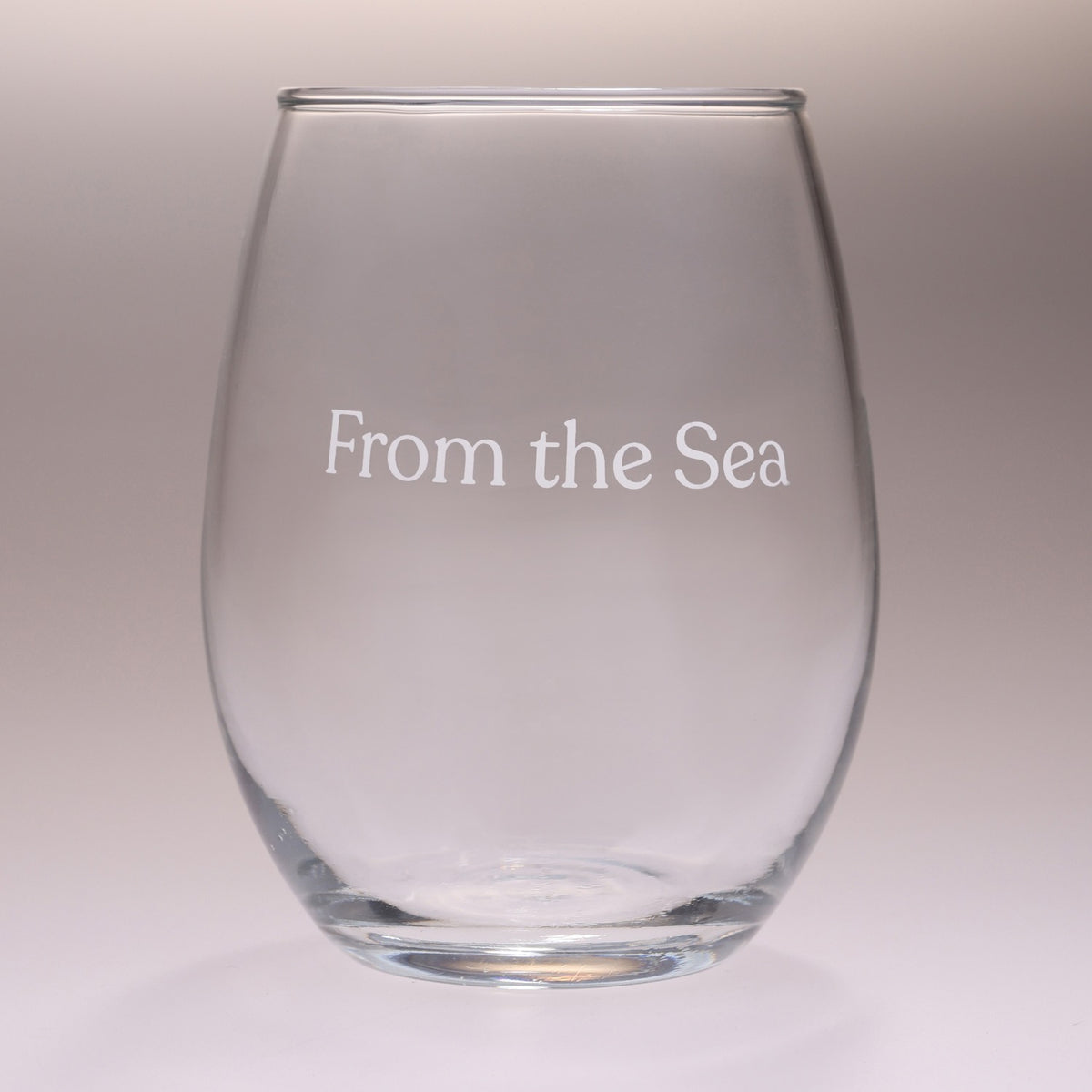 A clear stemless wine glass with the text 'From the Sea' printed in white on the front, set against a neutral gray gradient background.