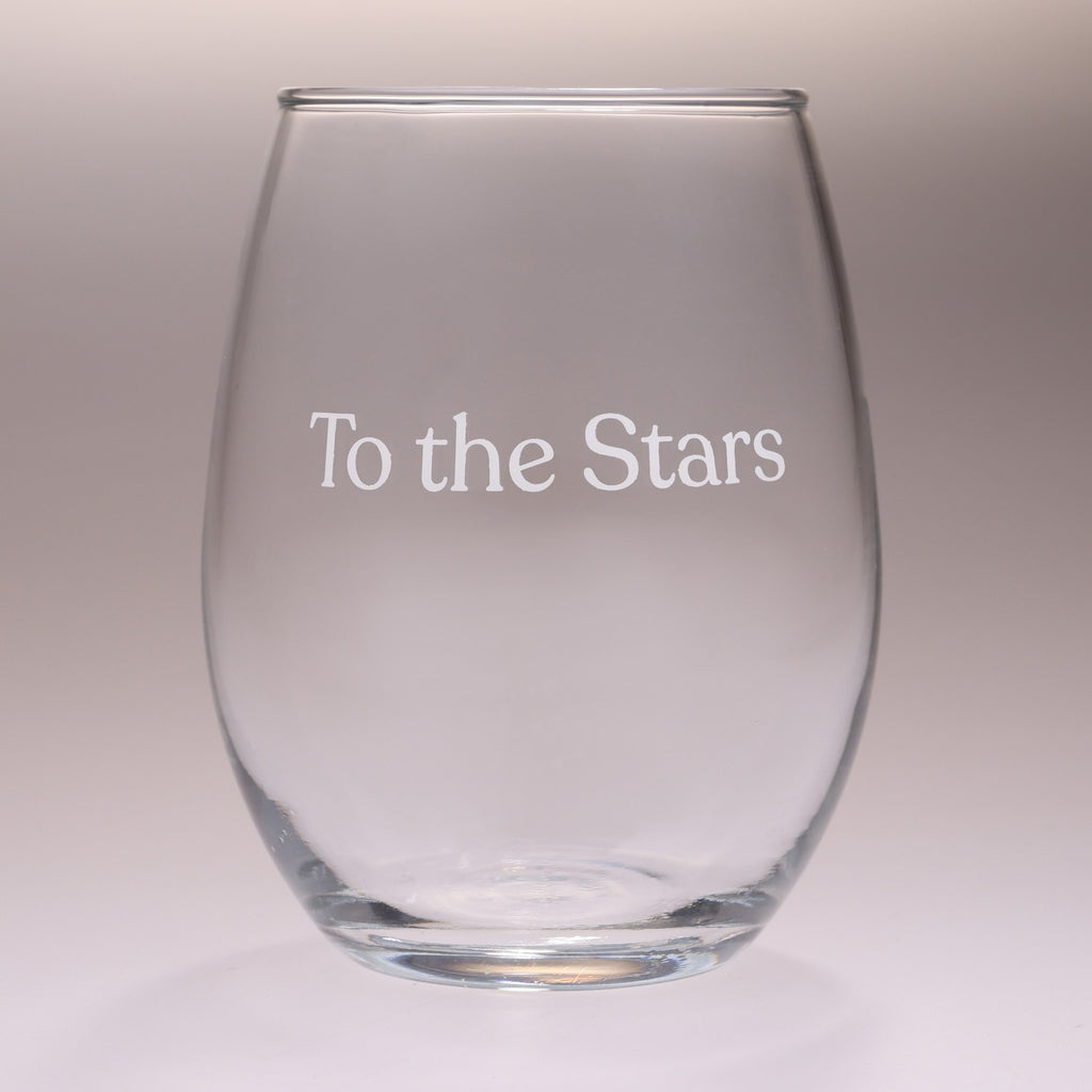 A clear stemless wine glass with the phrase 'To the Stars' printed in white serif lettering on the front, set against a neutral gray gradient background.
