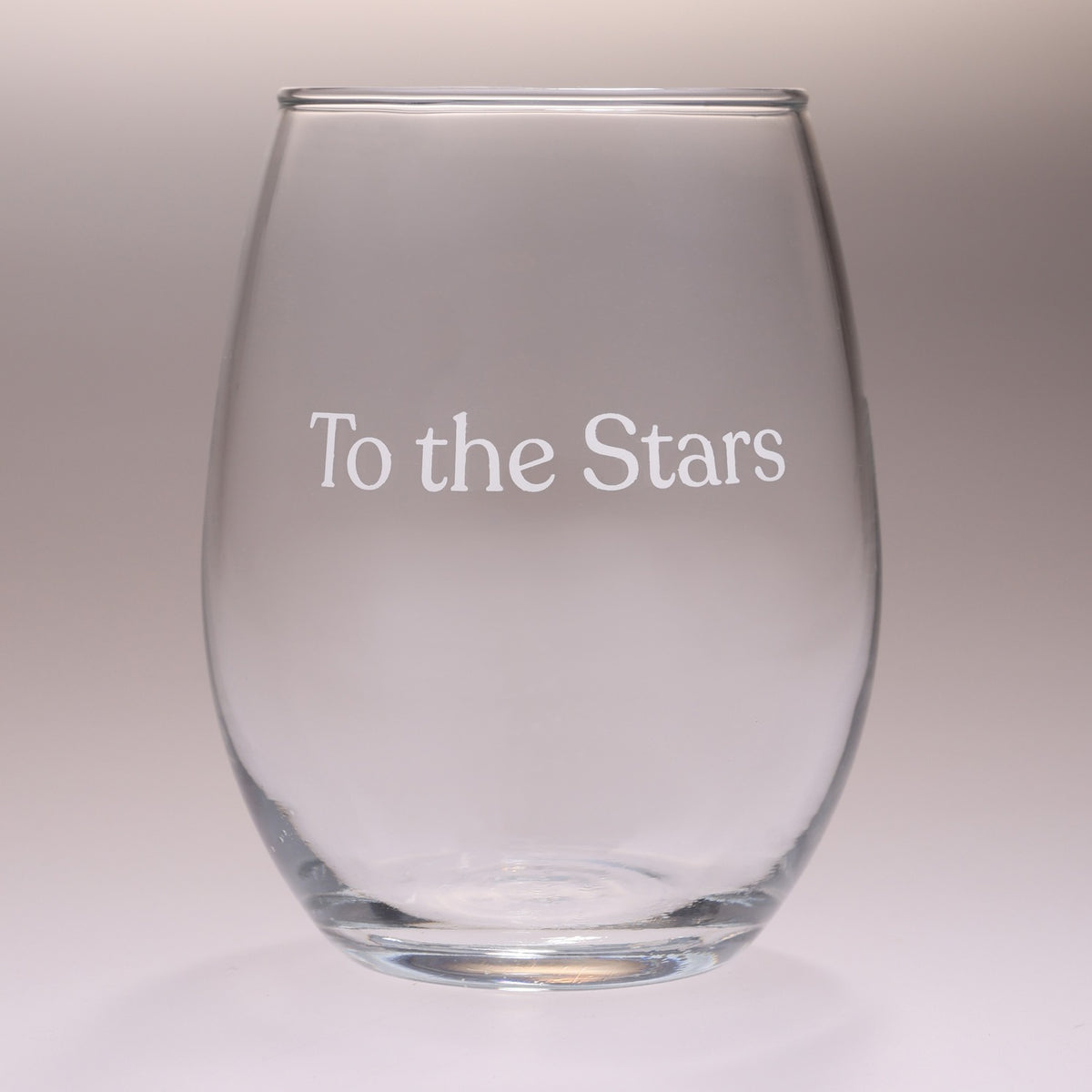 A clear stemless wine glass with the phrase 'To the Stars' printed in white serif lettering on the front, set against a neutral gray gradient background.
