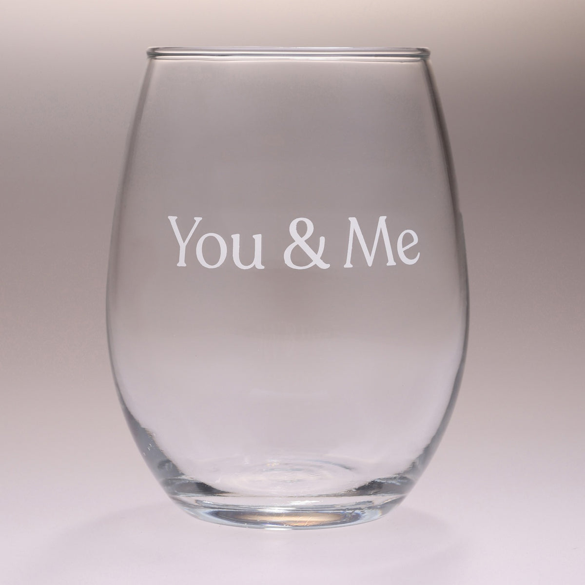 A clear stemless wine glass with the words 'You & Me' printed in white serif lettering on the front, set against a soft gray gradient background.