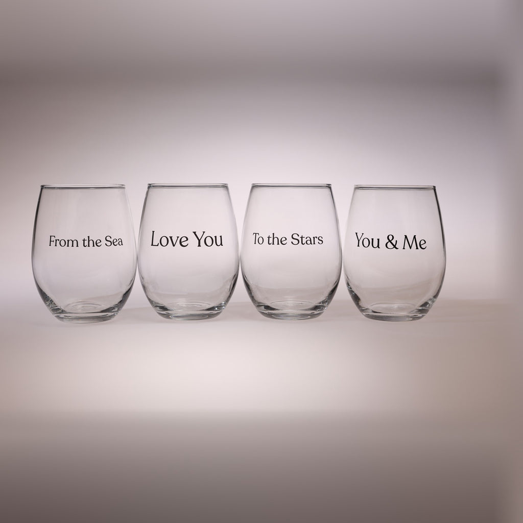 Four clear stemless wine glasses arranged in a row on a light gray gradient background. Each glass is printed with black text reading a different phrase: 'From the Sea,' 'Love You,' 'To the Stars,' and 'You & Me.