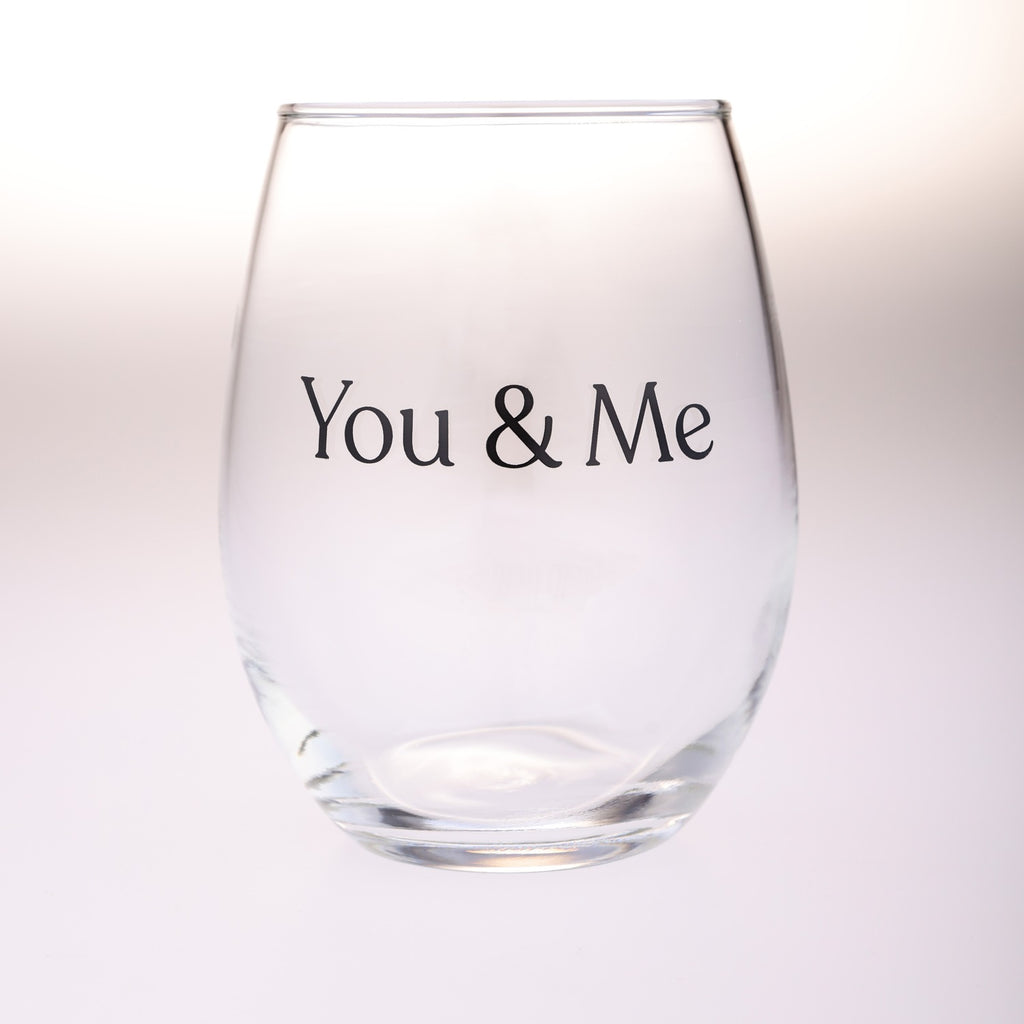 A clear stemless wine glass with the words 'You & Me' printed in black serif font on the front, shown against a soft white background.