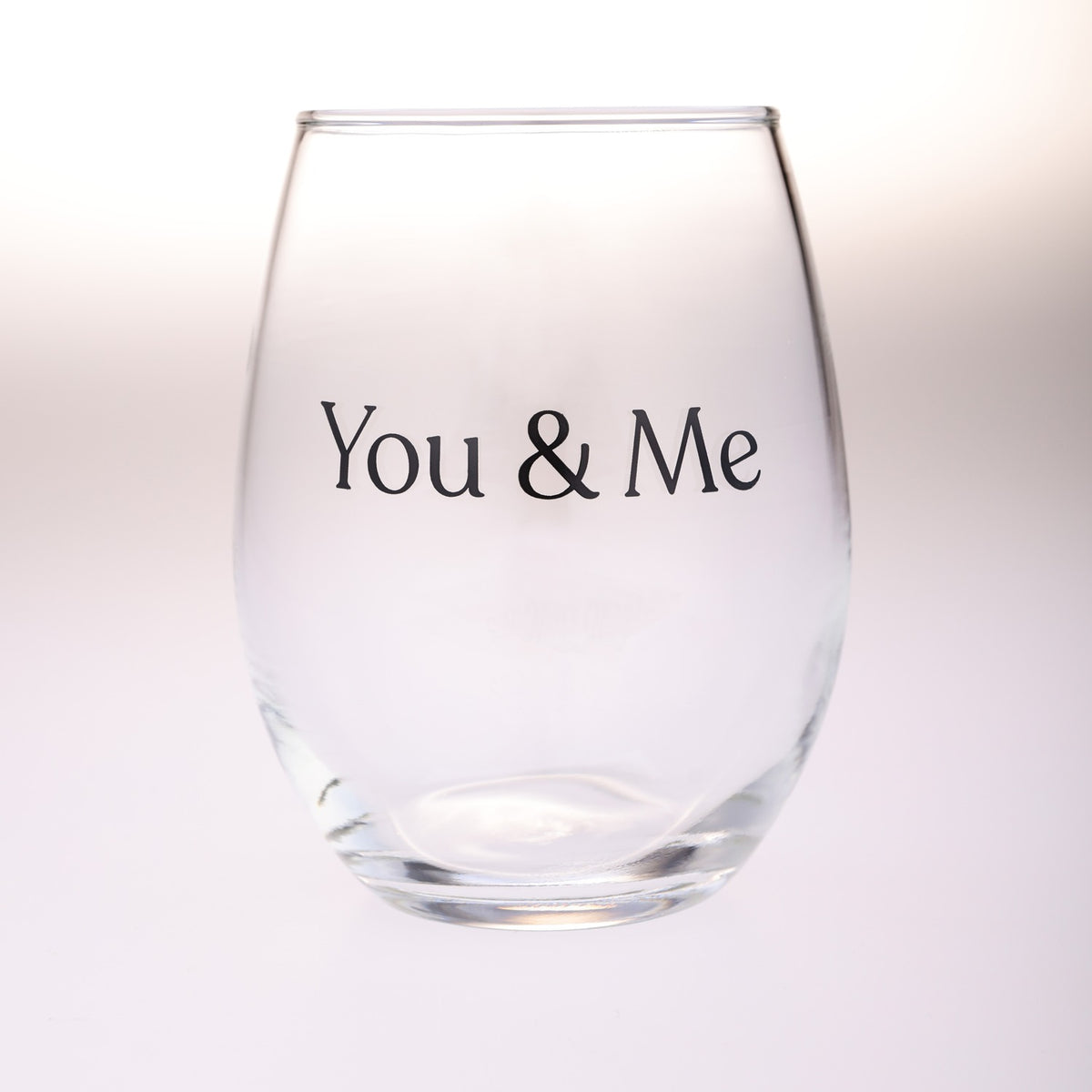 A clear stemless wine glass with the words 'You & Me' printed in black serif font on the front, shown against a soft white background.