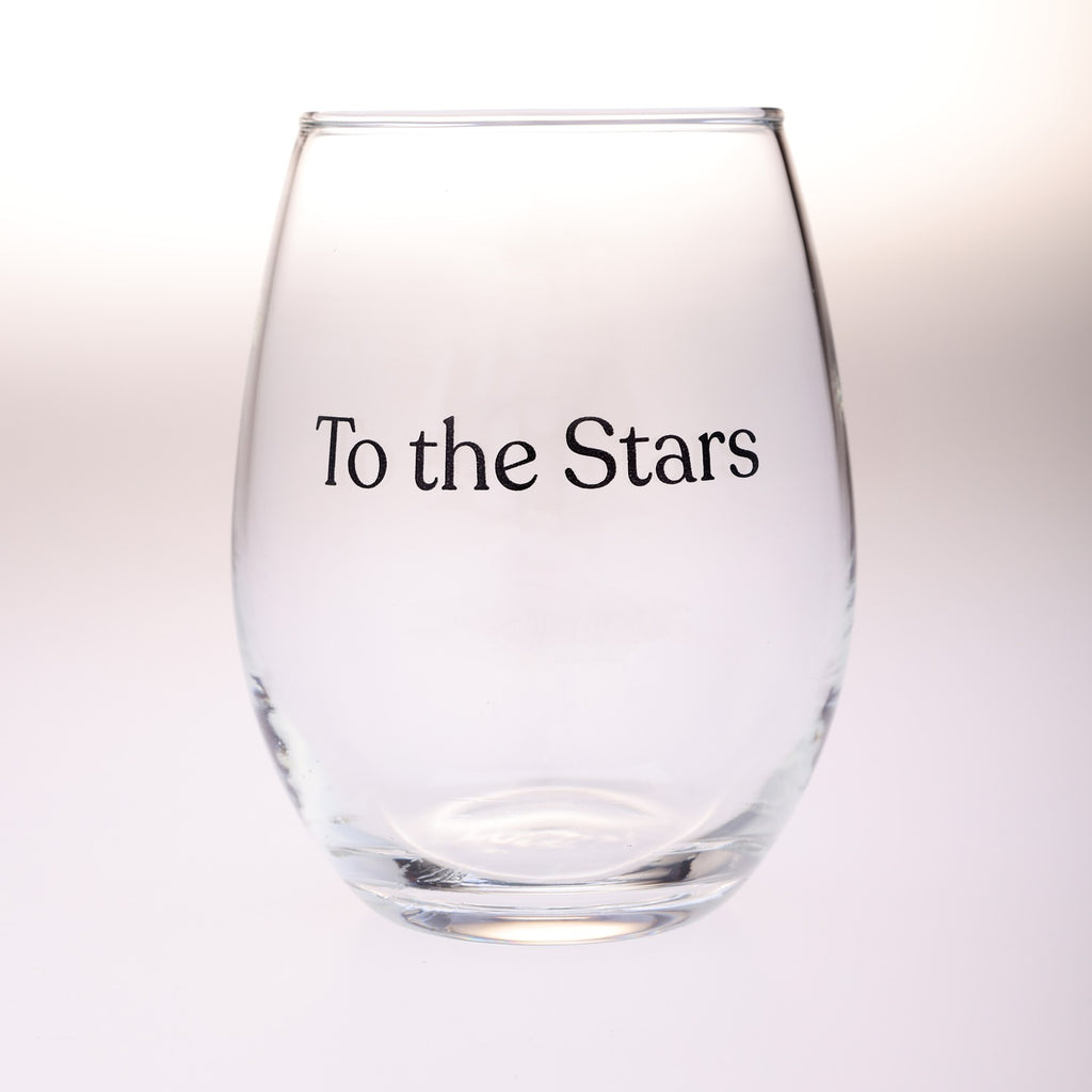 A clear stemless wine glass with the phrase 'To the Stars' printed in black serif text on the front, set against a soft white background.