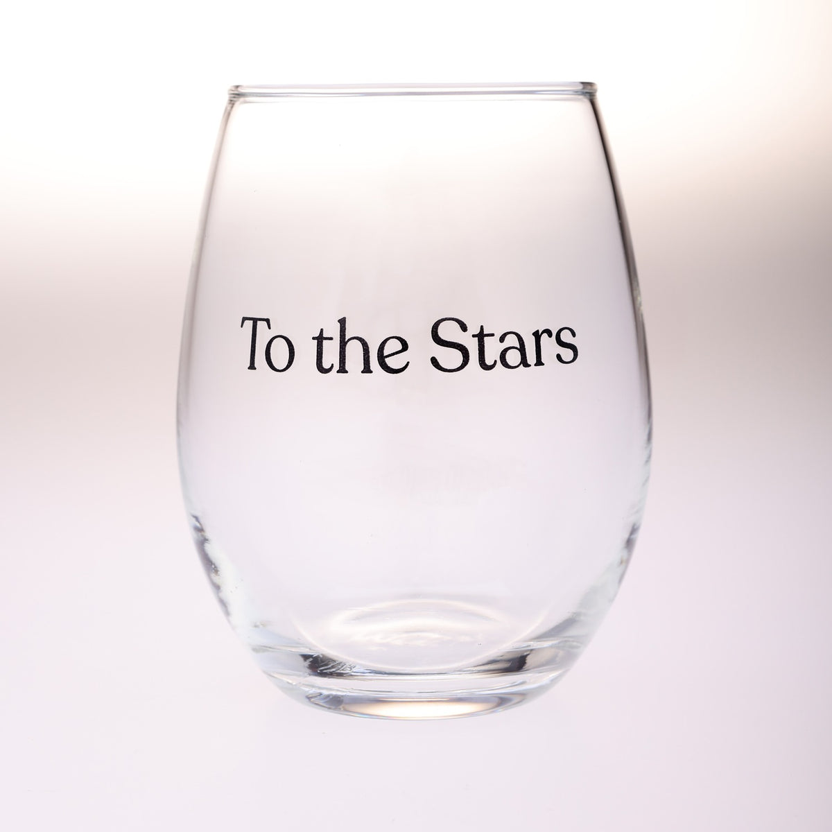 A clear stemless wine glass with the phrase 'To the Stars' printed in black serif text on the front, set against a soft white background.