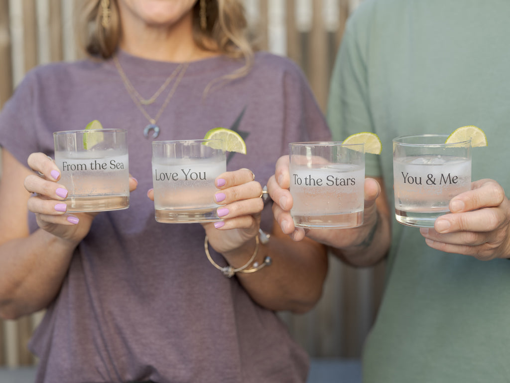 Four clear personalized rocks glasses held by the co-founders. The glasses are printed with romantic phrases: 'From the Sea', 'Love You', 'To the Stars', and 'You & Me'.