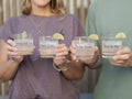 Four clear personalized rocks glasses held by the co-founders. The glasses are printed with romantic phrases: 'From the Sea', 'Love You', 'To the Stars', and 'You & Me'.